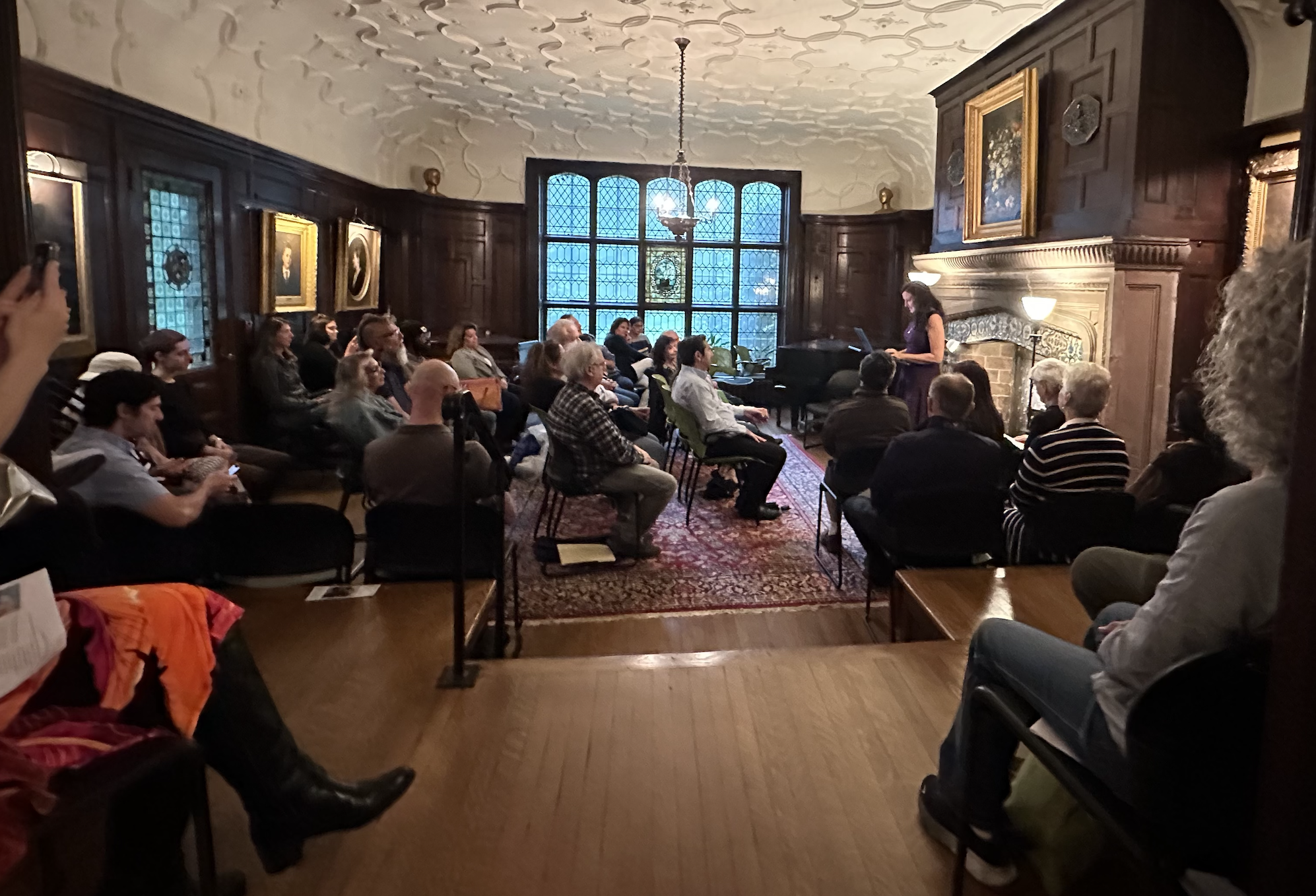 Poet reads in front of a grand fireplace to a packed audience. 