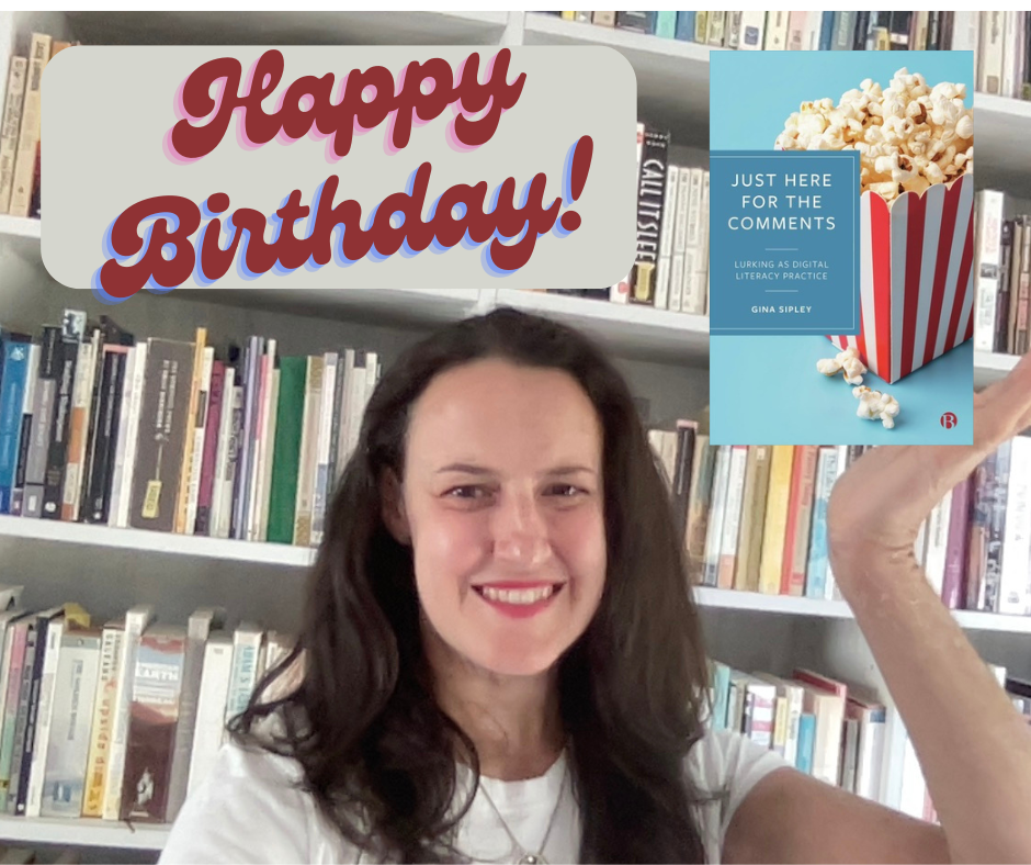 Gina Sipley stands in front of a book shelf and holds a copy of Just Here For The Comments below a Happy Birthday sign