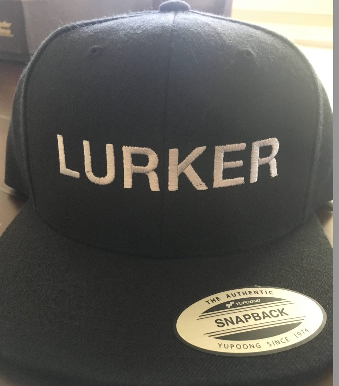 Black baseball cap with the word Lurker embroidered in white. 