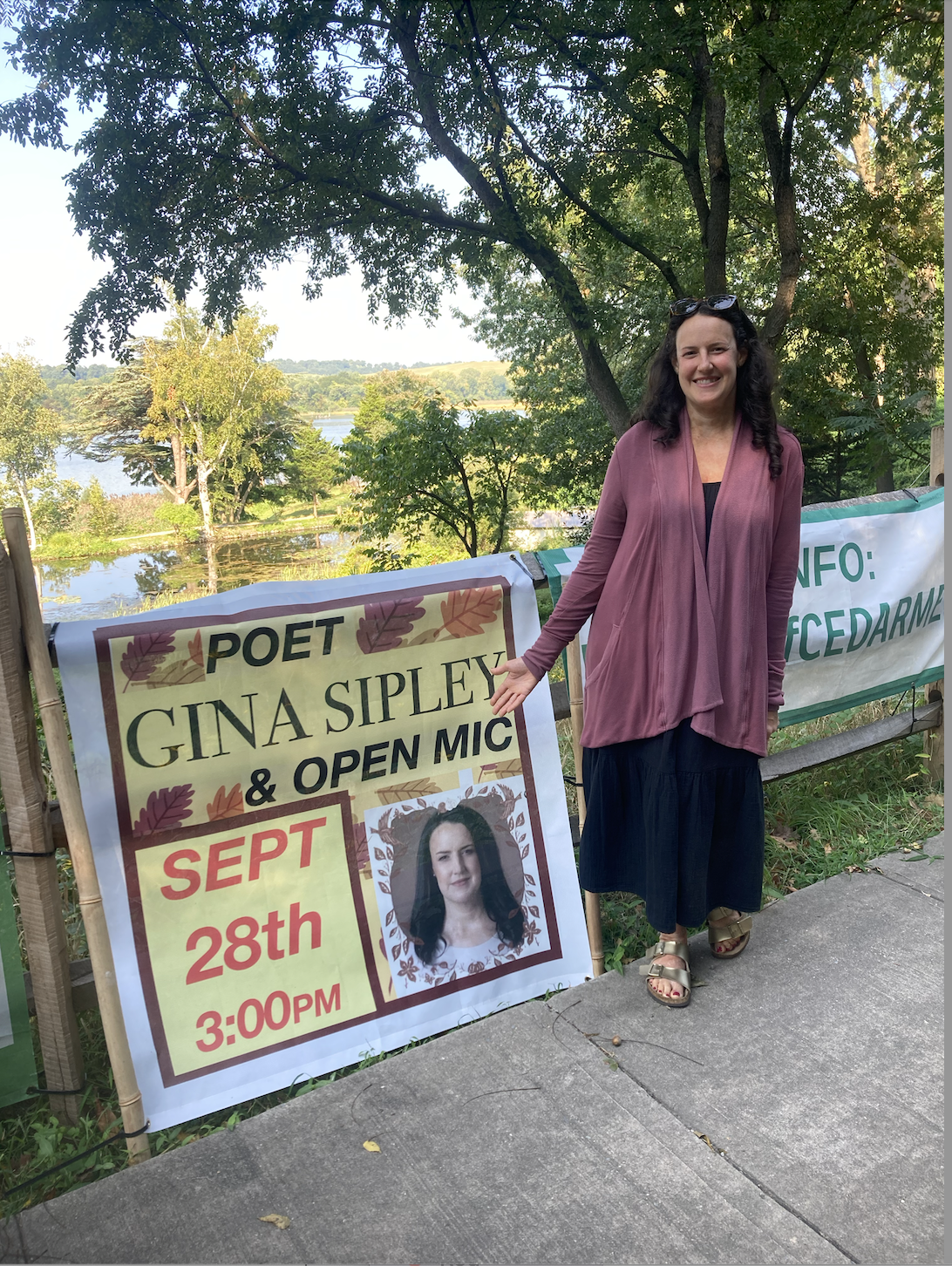 Woman stands next to a sign advertising a Poetry Open Mic at Cedarmere. 