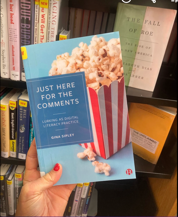 A hand holds a copy of Just Here For the Comments in front of a library book shelf. 